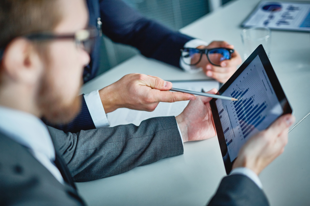 two businessmen in suits both looking at a tablet with one pointing at tablet with a pencil and holding a pair of glasses in his other hand. 