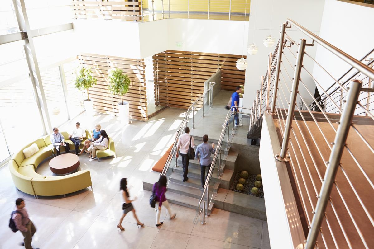 Modern office lobby with people walking and chatting. Features a central staircase, wood accents, circular seating area, and large windows. Bright and inviting.
