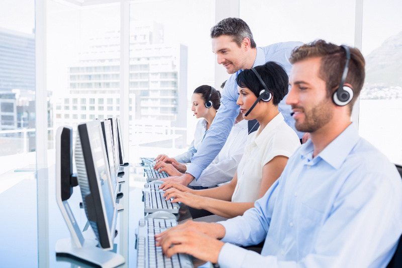 Office team in headsets works at computers in a bright office. A manager leans in, assisting a colleague.