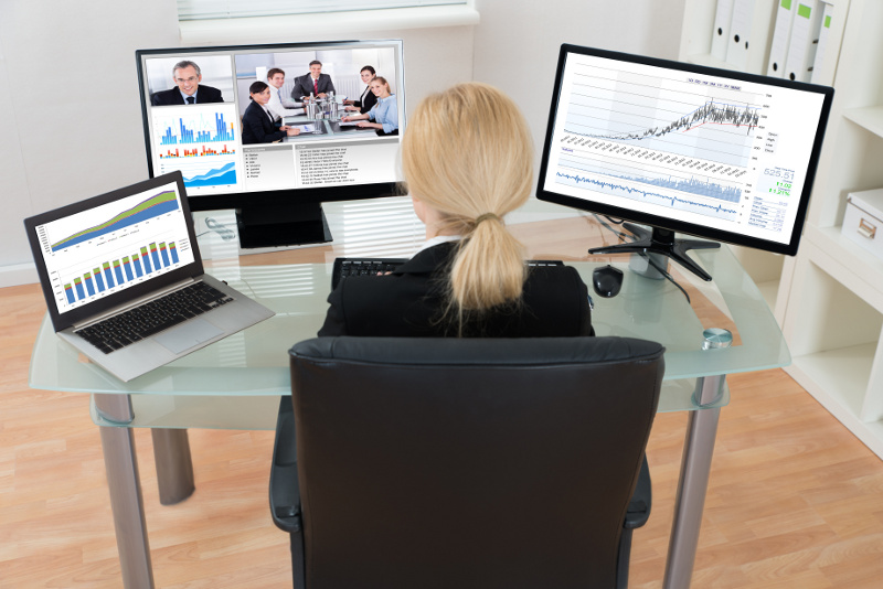 A woman sits at a glass desk with three monitors showing charts and a video conference. 